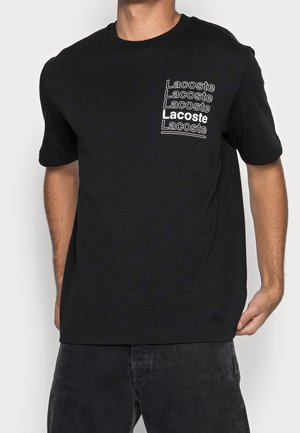 Personne portant un t-shirt noir avec le texte blanc répété "Lacoste" sur le haut à gauche de la poitrine, associé à un pantalon sombre.