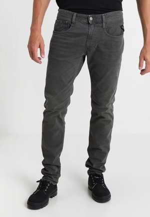 Mann trägt schlanke, dunkelgraue Jeans und schwarze Schnürstiefel, steht vor einem schlichten hellen Hintergrund.