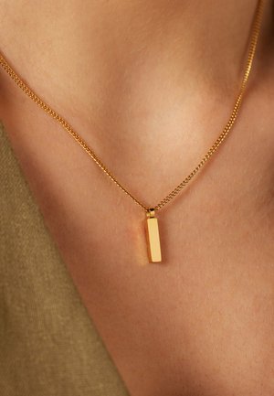 Goldene Halskette mit vertikalem rechteckigem Anhänger, getragen auf nackter Haut über einem beigen Stoffkleidungsstück.