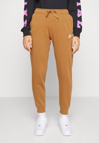 Nike joggers i en tan-färg, med dragsko i midjan, avsmalnande ben och en vit logotyp. Kombinerat med vita sneakers och en svart långärmad topp.