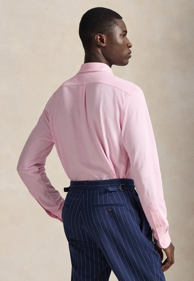 Light pink long-sleeve shirt with a button-down collar. Back pleat detail, paired with navy pinstriped trousers, featuring a belt and back pocket.