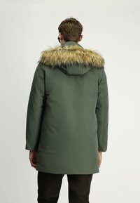 Mężczyzna w długim zielonym zimowym płaszczu z kapturem wykończonym futrem, pokazany z tyłu na jednolitym tle.