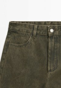 Spodnie dżinsowe w oliwkowej zieleni z przednią kieszenią, szlufką na pasek, metalowym zapięciem na guzik oraz widocznym przeszyciem na białym tle.