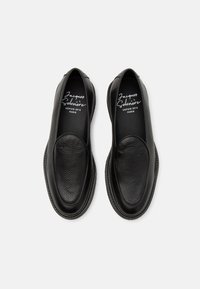 Black leather slip-on shoes with a textured finish, rounded toe, and contrasting stitching along the edge. Features a logo on the insole.
