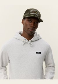 Sudadera de capucha gris con un parche de logo negro y cordones, combinada con una gorra de pana verde oscura con texto y detalles bordados.
