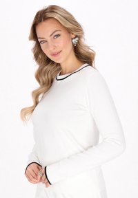Cămașă albă cu mâneci lungi, cu bordură neagră la gulerul rotund și la manșete; model care își ține mâinile împreunate, purtând accesorii îndrăznețe.