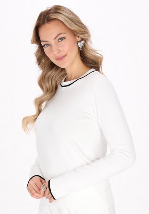 Chemise blanche à manches longues avec des bordures noires autour du col rond et des poignets ; modèle les mains jointes, portant des accessoires audacieux.