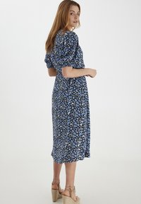 Blumenkleid in dunklem Marineblau mit blauen und grünen Blumen. Kurze puffärmel, tailliert und mit einem schwingenden Rock. Getragen mit hellbraunen Espadrilles mit Keilabsatz.