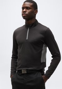 Άνδρας που φοράει μαύρο μακρυμάνικο πουκάμισο με φερμουάρ στο ένα τέταρτο και γκρι πλευρικές λωρίδες, καθώς και μαύρο παντελόνι, στέκεται με τα χέρια στις τσέπες.