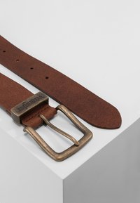 Brauner Leder-Gürtel mit glatter Oberfläche, ausgestattet mit einer bronzefarbenen Metallschnalle und graviertem Logodetail, auf einer hellgrauen Oberfläche platziert.