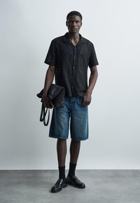 Mężczyzna ubrany w czarną koronkową koszulę z krótkim rękawem, dżinsowe szorty, czarne skarpetki i mokasyny, trzymający czarną złożoną torbę na jednolitym szarym tle.