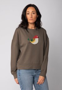 Vrouw met donker haar in een bruine sweatshirt met een geometrisch ontwerp van een rode, groene, gele en beige vogel, in combinatie met lichtblauwe jeans.