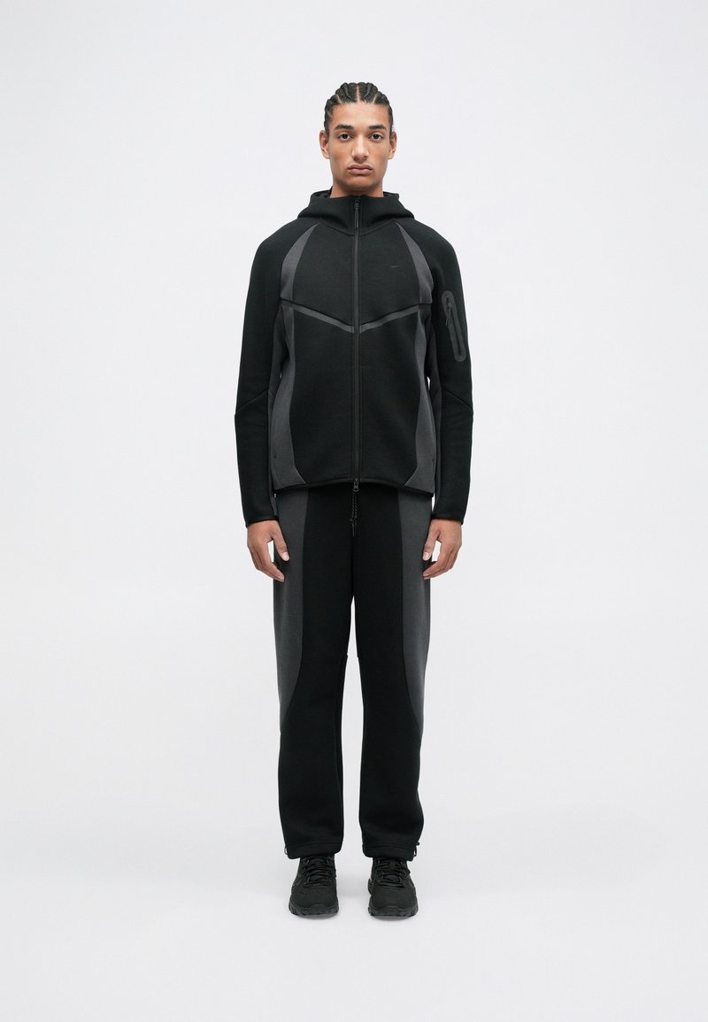 Černý sportovní outfit skládající se z mikiny na zip s kapucí a texturovanými akcenty a odpovídajících přiléhavých kalhot. Elegantní design s kontrastními šedými detaily.