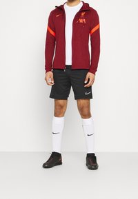 Bordeauxfarbene Sportjacke mit orangefarbenen Akzenten, mit Reißverschlusstasche, kombiniert mit schwarzen Shorts, weißen Socken und schwarzen Fußballschuhen mit roten Details.