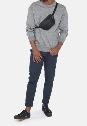 Man draagt een grijze trui, marineblauwe broek, zwarte sneakers en een zwarte schoudertas tegen een effen witte achtergrond.