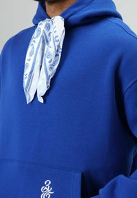 Blauwe hoodie met een voorzak, voorzien van een wit bandana met patroon, gebonden rond de hals. Zachte stof en een casual ontwerp.