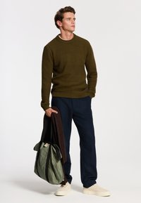 Mann in olivgrünem Pullover und navyblauer Hose hält eine grüne Tragetasche und einen braunen Schal und schaut zur Seite vor einem schlichten weißen Hintergrund.