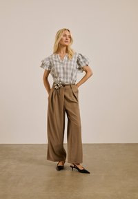 Femme portant un pantalon beige à jambes larges avec une ceinture florale et un chemisier blanc à carreaux avec des manches à volants, debout sur un fond uni.
