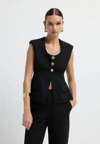 Donna con capelli lisci e raccolti indossa un blazer senza maniche nero con bottoni dorati e pantaloni coordinati, accessoriata con orecchini a cerchio dorati e un bracciale.