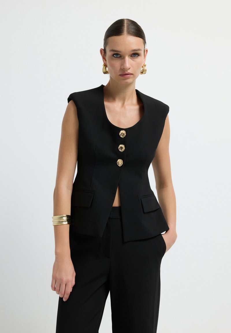 Donna con capelli lisci e raccolti indossa un blazer senza maniche nero con bottoni dorati e pantaloni coordinati, accessoriata con orecchini a cerchio dorati e un bracciale.