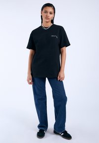 Camiseta de algodón negra de manga corta con un logo en el pecho, combinada con unos pantalones vaqueros de pierna ancha de mezclilla azul y zapatillas negras con detalles en blanco.