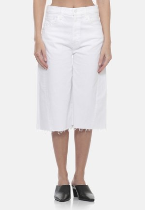 Donna che indossa jeans bianchi a gamba larga e corta con orli sfrangiati e scarpe nere con tacco slip-on, in piedi davanti a uno sfondo bianco.