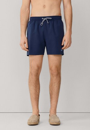 Hombre con pantalones cortos de baño azul marino con cordón blanco y zapatos beige sin cordones, de pie contra un fondo claro y liso.