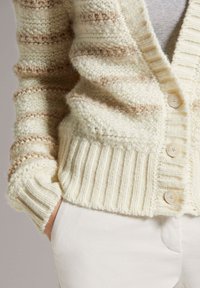 Crème cardigan met horizontale beige strepen, ribgebreide manchetten en een gestructureerde afwerking. Bevat subtiele knoopaccenten en heeft een ontspannen pasvorm.