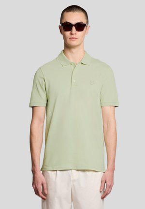 Jonge man met een lichtgroen poloshirt met korte mouwen, crèmekleurige broek en donkere zonnebril, staand tegen een effen grijze achtergrond.