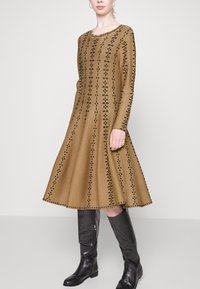 Brązowa dzianinowa sukienka z czarnymi kwiatowymi wzorami, długimi rękawami i rozkloszowanym dołem; w połączeniu z czarnymi skórzanymi kozakami do kolan.
