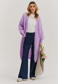 Fialový mohairový kardigán cez bielu košeľu, kombinovaný s tmavomodrými rozšírenými džínsami a svetlosivými semišovými topánkami. Ruka drží sivú tašku s kvetmi.