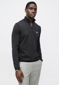 Camisola preta com fecho de correr, textura canelada e logótipo branco "BOSS" no peito, combinada com calças cinza claro às riscas finas.