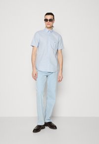 Homme portant une chemise bleue claire à manches courtes boutonnée, un jean bleu clair, des chaussures noires et des lunettes de soleil foncées, debout devant un fond blanc uni.