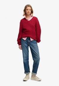 Une jeune femme se tient debout, portant un pull rouge en maille torsadée par-dessus une chemise rose à col, un jean bleu retroussé et des baskets montantes blanches.