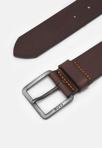 Brun skinnbelte med en strukturert finish, med et metallspenne som har en slitt look. Oransje sømdetalj nær spennen.