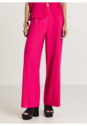 Femme portant un pantalon large rose vif, des talons à plateforme ouverts noirs et un haut assorti rose vif, avec des bracelets dorés et roses au poignet gauche.