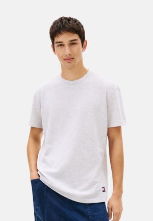 Joven con camiseta de manga corta gris claro y pantalones azul oscuro, de pie con una mano en el bolsillo contra un fondo blanco.