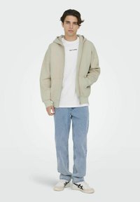 Lichtbeige ritsvest met een ruime pasvorm, gecombineerd met een witte ronde hals t-shirt, lichtblauwe jeans en witte sneakers.
