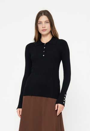 Femme portant un pull noir côtelé ajusté à manches longues avec des boutons sur la patte de boutonnage avant et aux poignets, associé à une jupe marron.