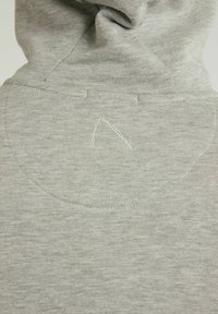Tejido de sudadera gris claro con costuras visibles y un sutil logo bordado en la parte posterior, cerca de la costura de la capucha.