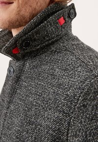 Abrigo gris texturizado con cuello alto, botones negros y detalles en rojo. La tela presenta un diseño jaspeado y un ajuste estructurado.