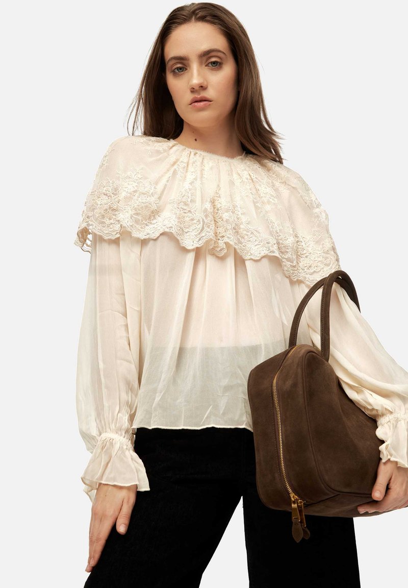 Cream blouse with lace trim, flowing design, long sleeves with gathered cuffs; paired with a brown suede bag featuring a zip closure.