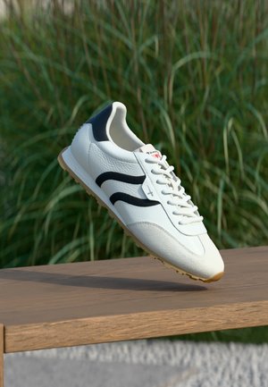 Biały skórzany sneaker z czarnymi akcentami i teksturowanymi detalami, okrągły nosek oraz białe sznurowadła. Podeszwa gumowa, unosi się na drewnianej powierzchni.