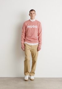 Ružová mikina s logom "HUGO", bielou košeľou s golierom pod ňou, béžovými nohavicami a bielymi teniskami. Jednoduchý, neformálny dizajn, minimálna textúra.