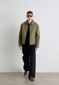 Veste vert olive avec fermeture éclair et col, haut noir, pantalon large noir et chaussures marron. Le modèle porte des lunettes de soleil noires. Fond uni.