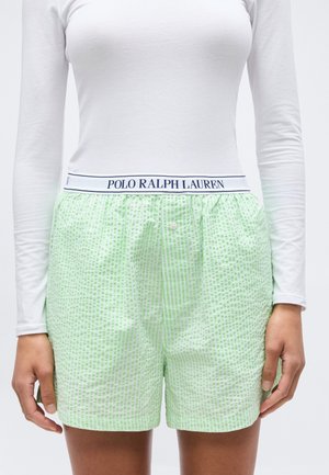 Kobieta ubrana w jasnziezielone teksturowane szorty oraz białą bluzkę z długim rękawem, z widocznym paskiem z logo Polo Ralph Lauren.