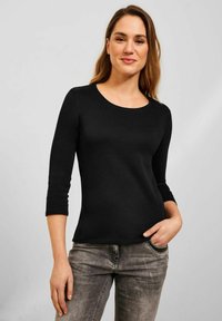 Schwarzes Langarmshirt mit rundem Ausschnitt, aus glattem Stoff, kombiniert mit ausgebleichten grauen Jeans. Das Shirt hat eine taillierte Passform.