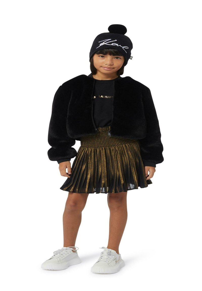 Veste en fausse fourrure noire courte, t-shirt noir avec inscription dorée, jupe plissée dorée et baskets blanches avec accents dorés. Bonnet pompon noir.