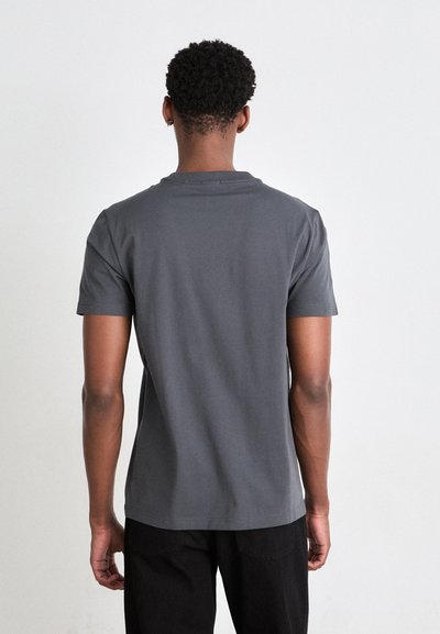 Homme aux cheveux courts et bouclés portant un t-shirt gris foncé uni à manches courtes et un pantalon noir, vu de dos sur fond blanc.