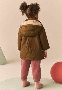 Brun hoodie-jacka med beige fleecefoder, rosa kordbyxor och rosa gummistövlar. Bakgrunden har gröna former och en röd sfär.